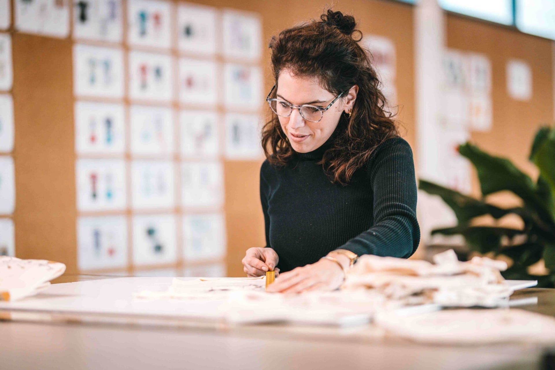 Ontwerper werkt met stof aan een werktafel, voor een wand met kledingontwerpen