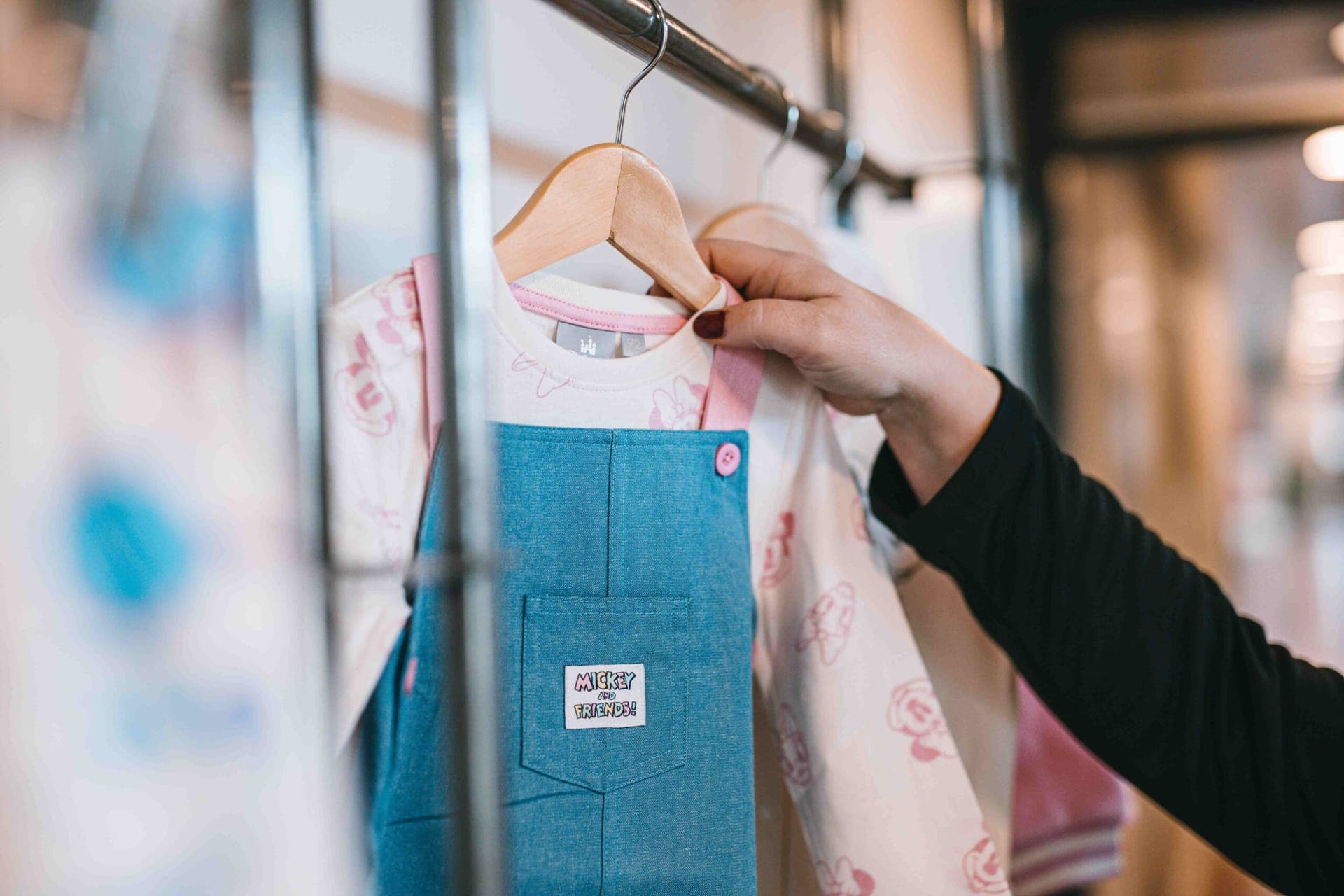 Close-up van een 'Mickey and Friends'-label op een denim tuinjurk, naast een top met Minnie Mouse-print